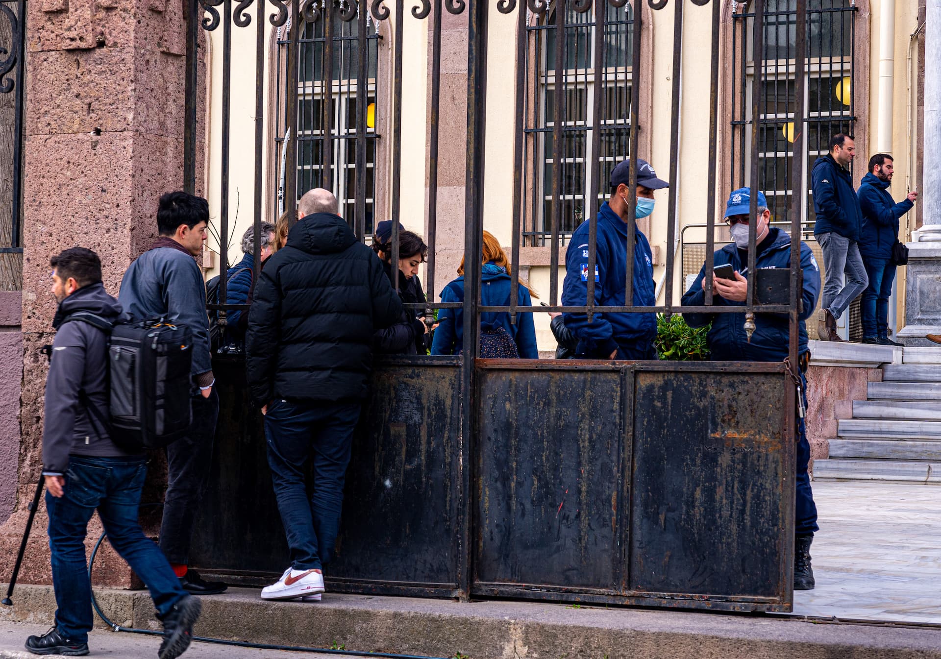 Seán stands and talks to someone through the bars of an iron gate. Police stand on the other side of the gate.